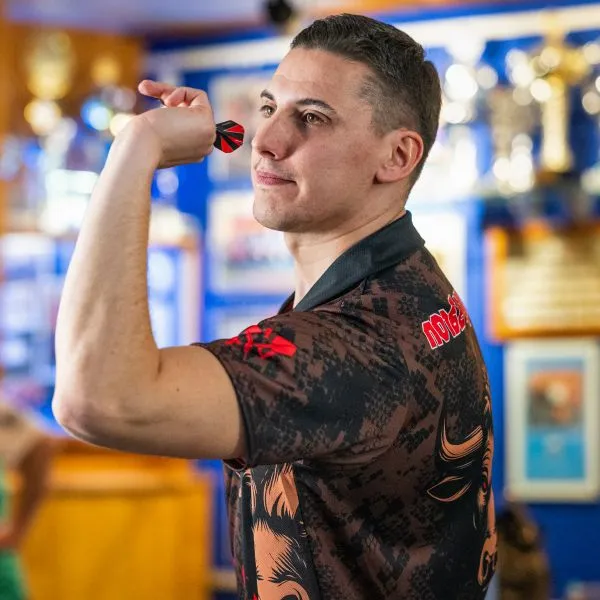 Un homme portant un maillot sportif à motifs avec des accents rouges tient un dart et vise, des trophées étant visibles en arrière-plan.