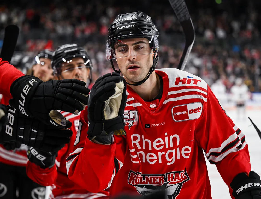 Un joueur de hockey porte un maillot rouge avec des détails blancs et noirs tout en interagissant avec un coéquipier.