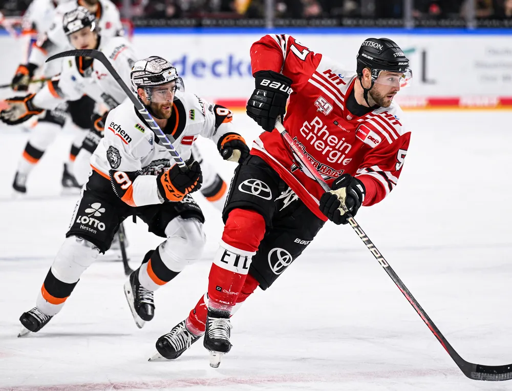 Un joueur de hockey sur glace en maillot rouge et pantalon noir patine sur la glace tout en étant poursuivi par un autre joueur.