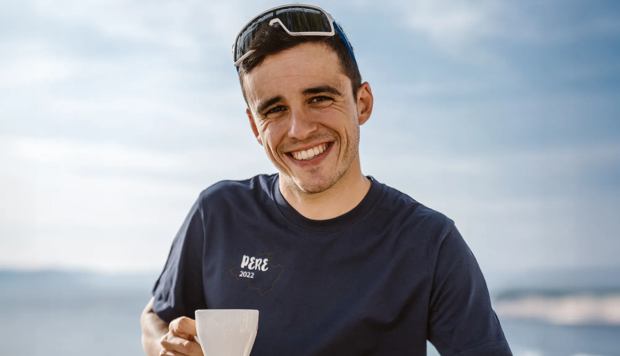 Un jeune homme sourit dans un t-shirt bleu avec le texte 'PERE 2022' tout en tenant une tasse, devant un ciel dégagé.
