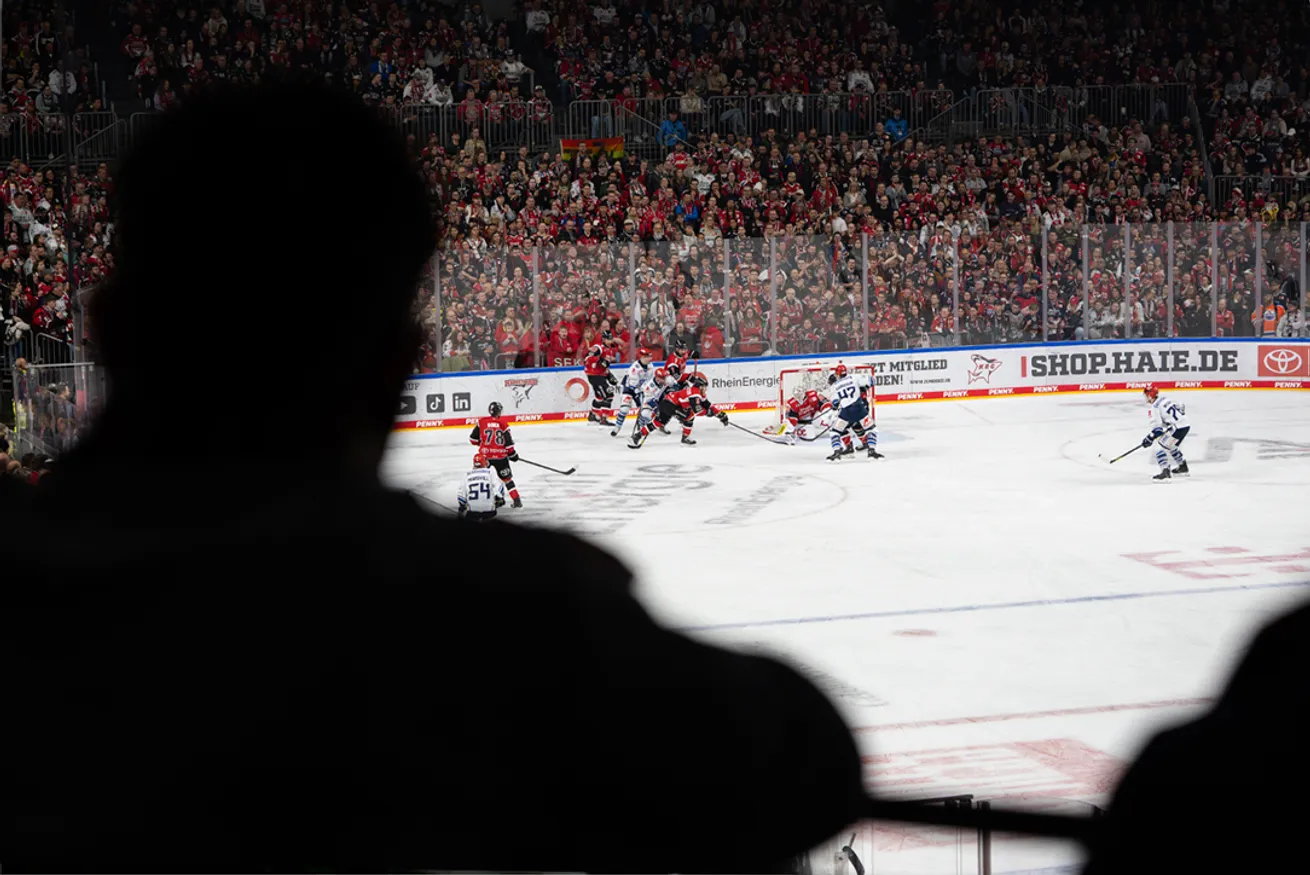 Du point de vue d'un spectateur, l'image montre un match de hockey sur glace avec des joueurs en maillots rouges et bleus devant une grande foule.