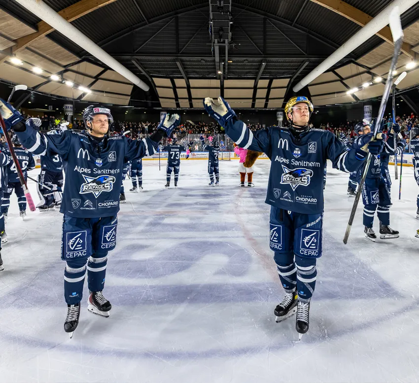 Des joueurs de hockey sur glace en maillots bleus avec des logos de sponsors célèbrent sur la glace, avec le public en arrière-plan.