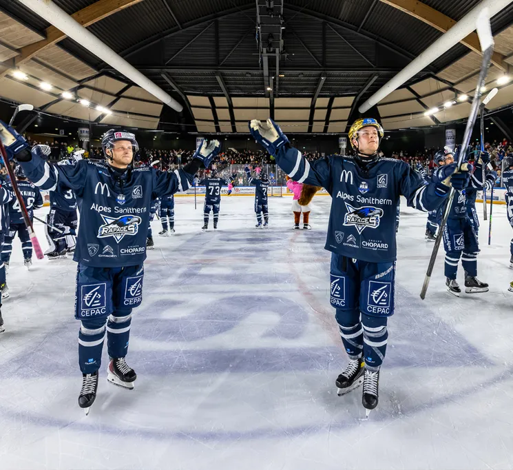 Des joueurs de hockey sur glace en maillots bleus avec des logos de sponsors célèbrent sur la glace, avec le public en arrière-plan.
