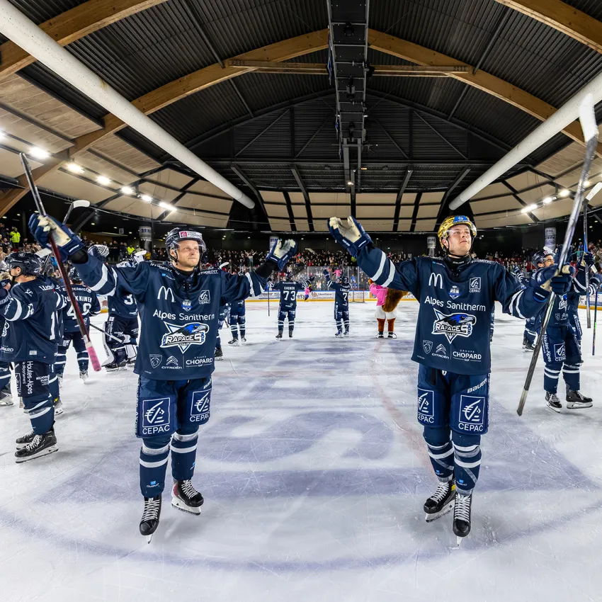 Deux joueurs de hockey sur glace en maillots bleus se tenant sur la glace, levant leurs bâtons tandis qu'une foule enjouée est visible en arrière-plan.