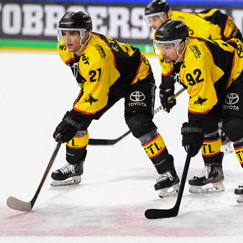 Deux joueurs de hockey sur glace en maillots jaunes et noirs se tiennent sur la glace, se préparant pour le match.