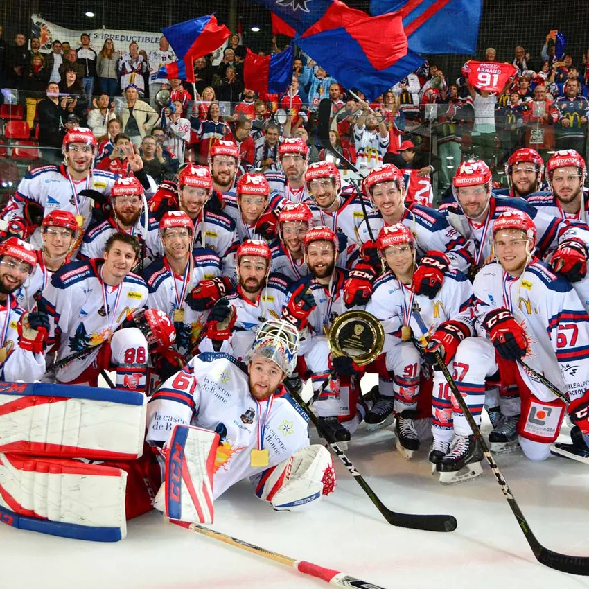 Une équipe de hockey pose fièrement sur la glace avec des médailles et un trophée pendant que des fans acclament en arrière-plan.