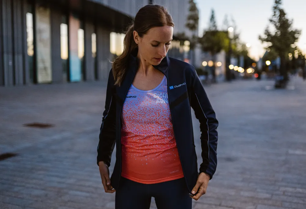 Une femme sportive porte une veste bleu foncé sur un débardeur sans manches dégradé bleu et orange, tandis qu'elle se tient dans un environnement urbain.