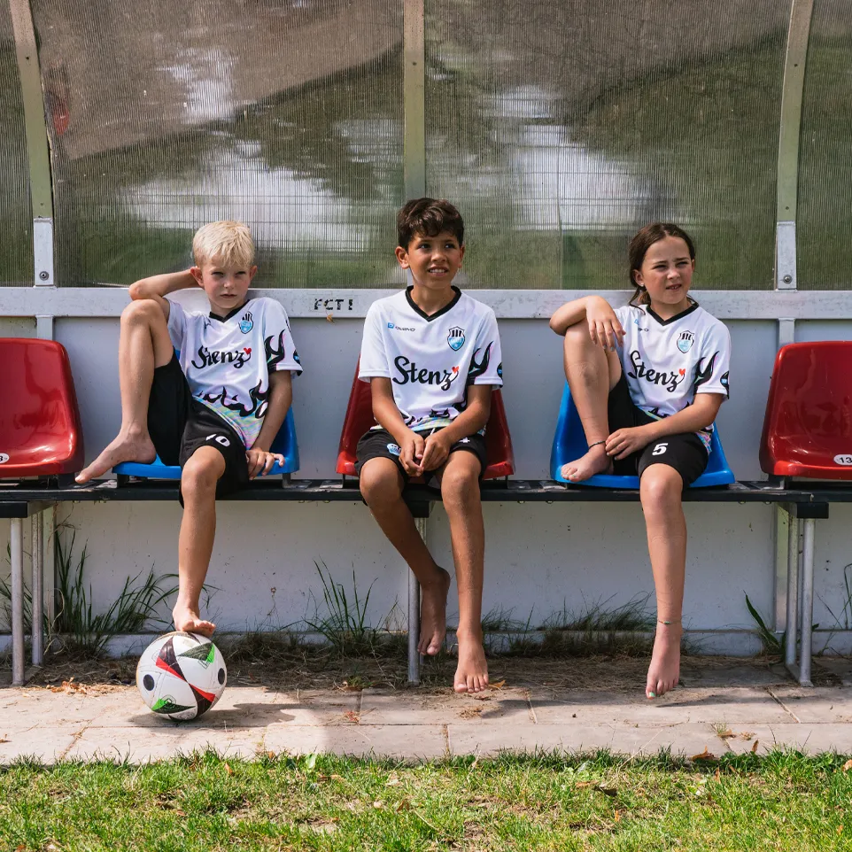 Trois enfants en maillots de football et shorts sont assis sur un banc au bord du terrain, avec un ballon de football devant eux.