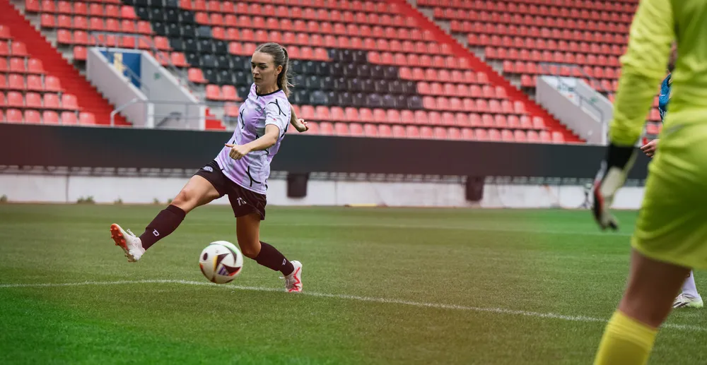Une joueuse en maillot de football violet et blanc frappe le ballon sur un terrain de stade, avec un gardien de but en équipement jaune en arrière-plan.
