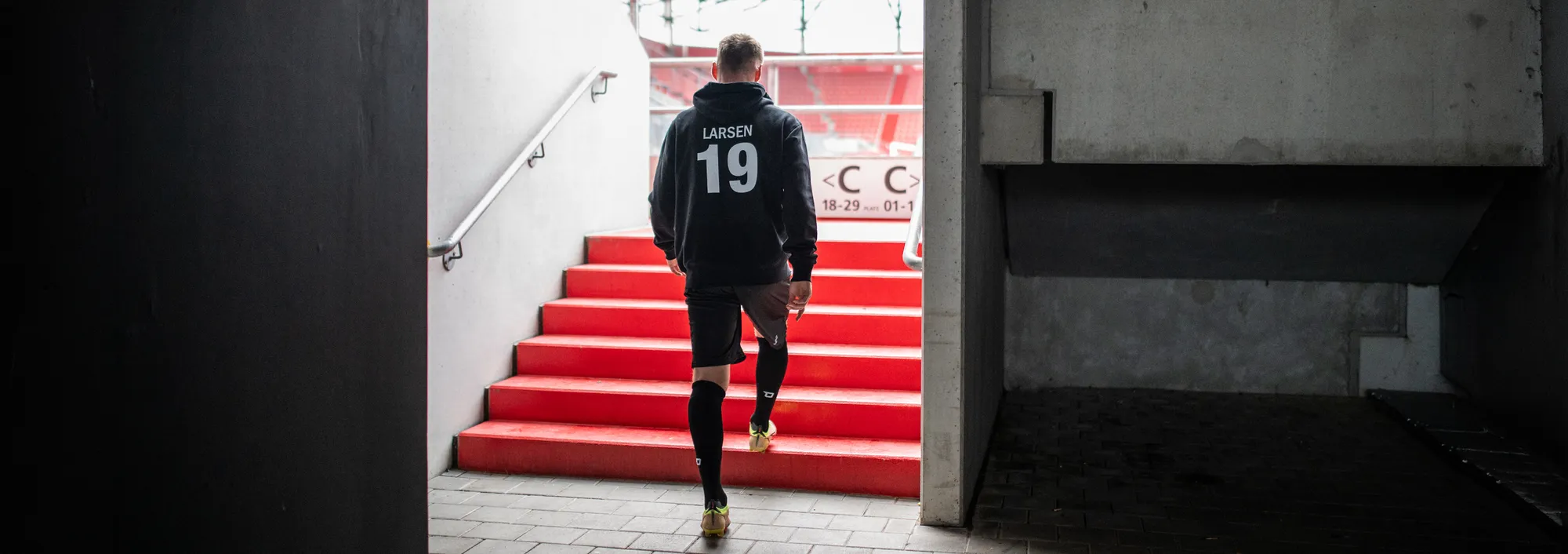 Un joueur portant un hoodie noir avec le numéro 19 descend des escaliers rouges en sortant d'un stade.