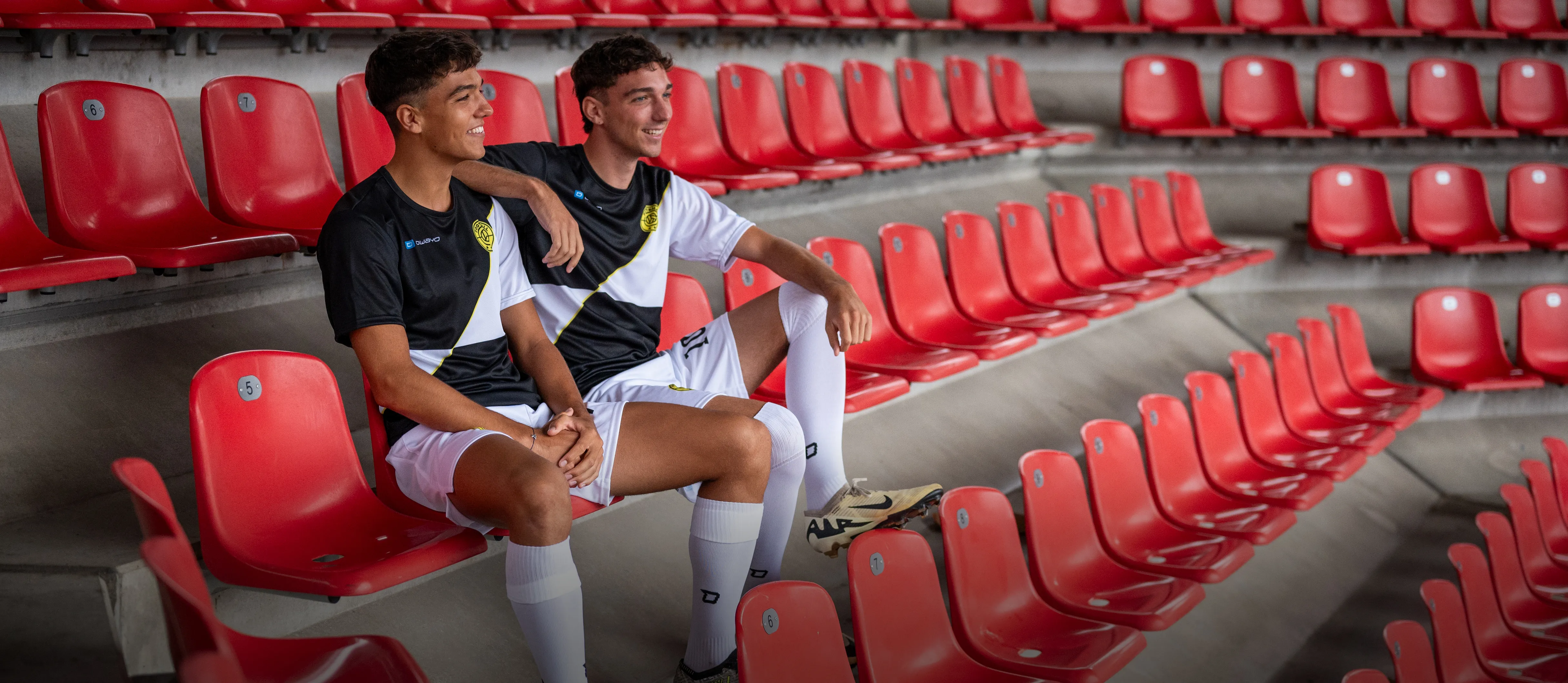 Deux jeunes hommes en tenue de football sportive sont assis décontractés sur des sièges de stade rouges.