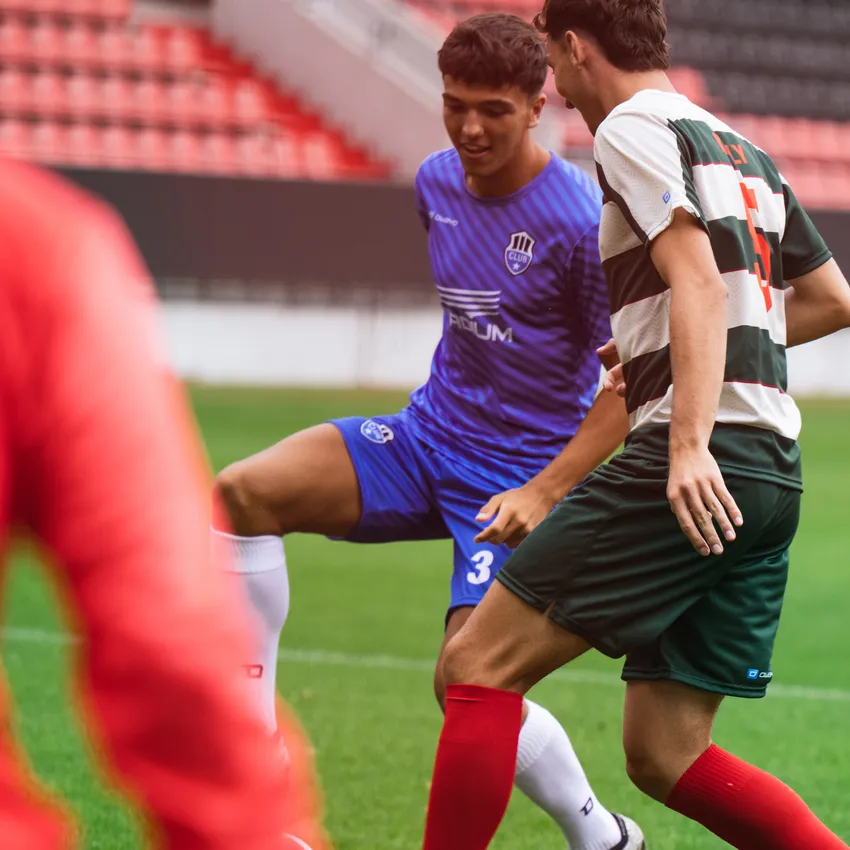 Deux joueurs de football en action sur le terrain, l'un portant un maillot bleu et l'autre un maillot rayé vert et rouge.