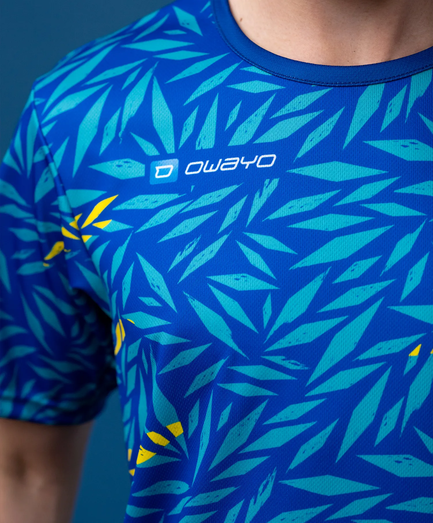 Un t-shirt sportif à manches courtes avec un motif bleu de type feuillage dans différentes nuances de vert et des accents jaunes.