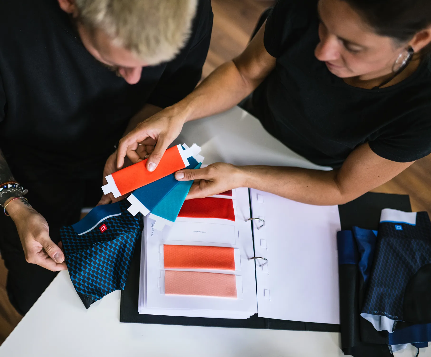 Deux personnes choisissent des couleurs pour des vêtements de sport personnalisés en examinant des échantillons de tissu et un nuancier.