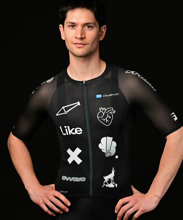Un jeune homme porte un haut de sport noir ajusté orné de différents logos et motifs blancs, posant devant un fond noir.