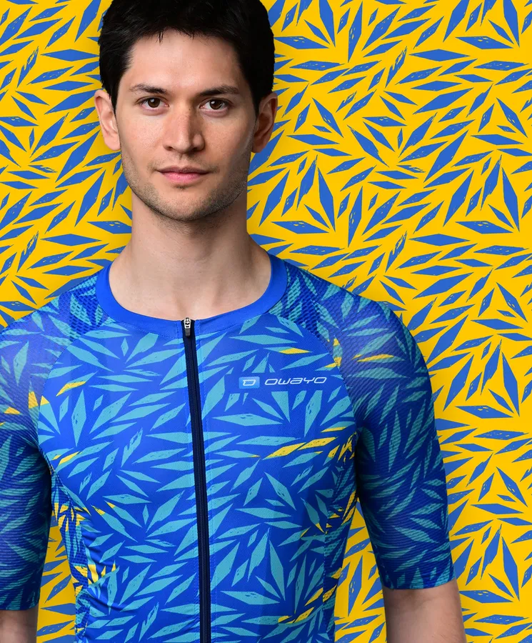 Un homme porte un maillot de sport bleu à manches courtes avec un motif de feuilles bleues et jaunes ressemblant à une plante tropicale, devant un fond jaune avec des motifs de feuilles bleues.