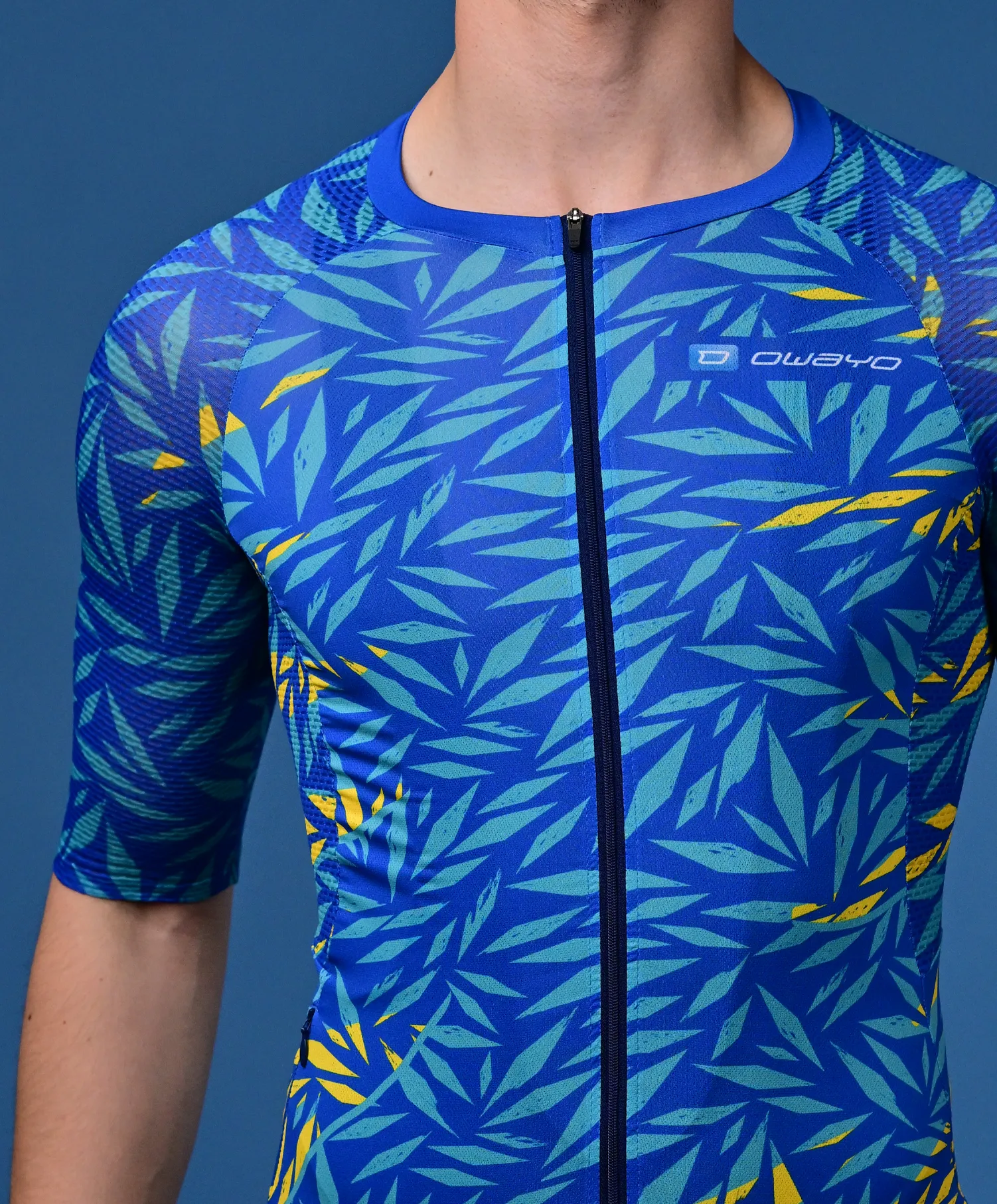 Un homme porte un maillot de sport bleu à manches courtes avec un motif de feuilles tropicales en turquoise et jaune et une fermeture éclair devant.