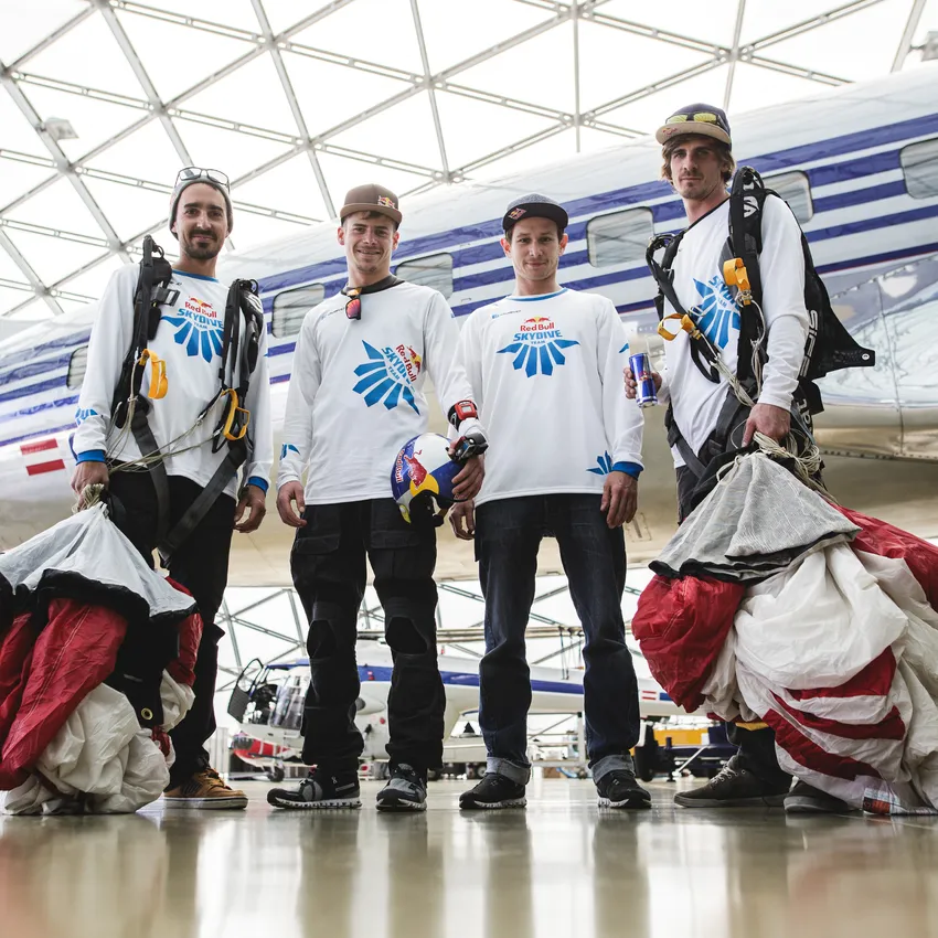 Quatre personnes en tenue sportive se tiennent dans un hangar moderne, prêtes pour un saut en parachute.