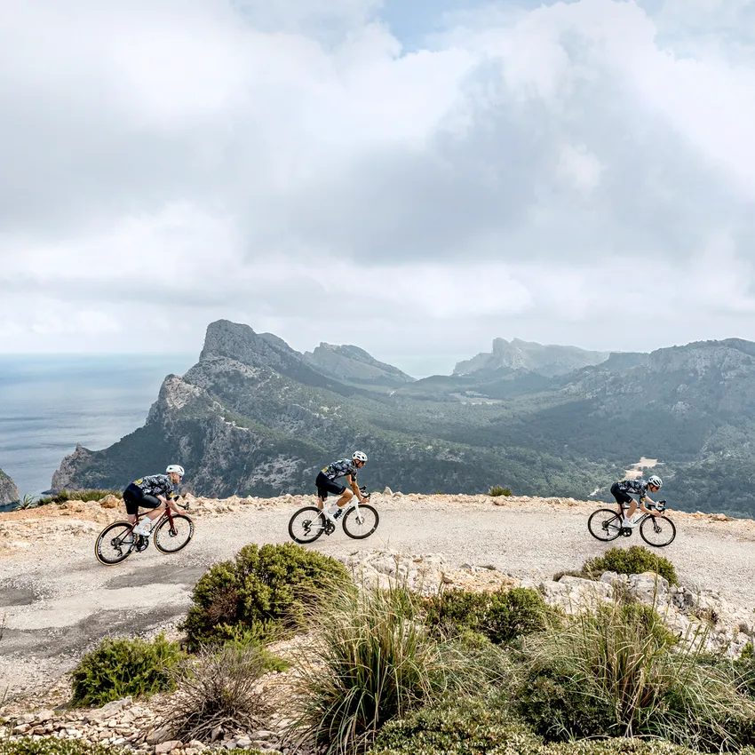 Trois cyclistes en tenue de sport roulent le long d'un chemin étroit avec vue sur un paysage côtier.