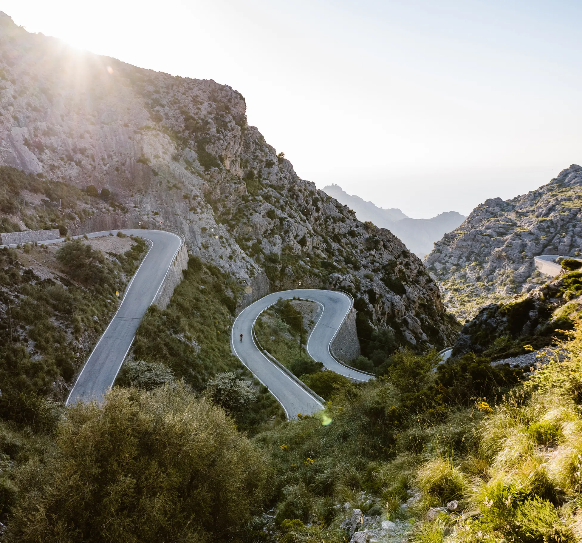 Une personne fait du vélo sur une route de montagne sinueuse qui serpente à travers un paysage rocheux, avec la lumière du soleil venant d'en haut.