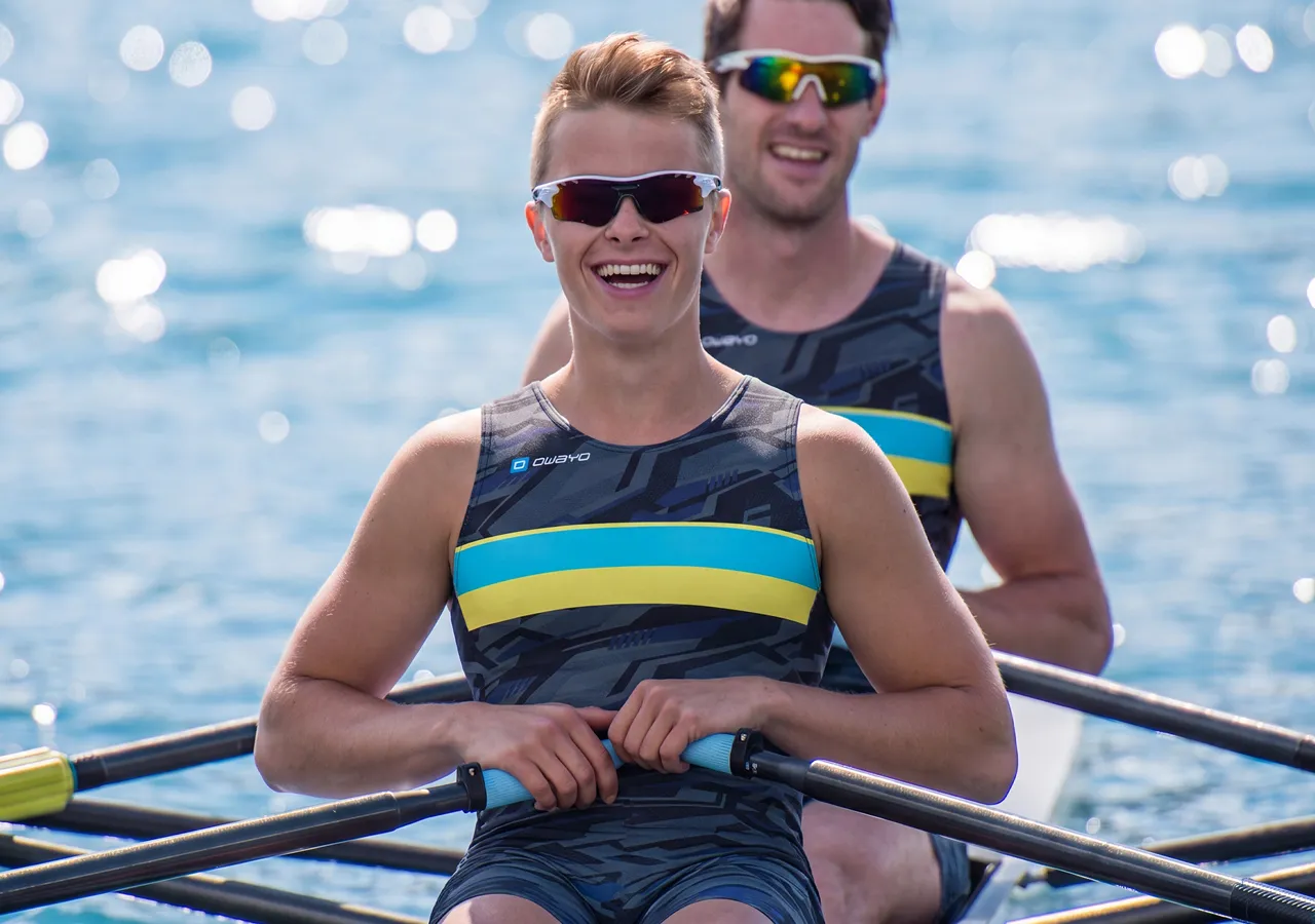 Deux athlètes en vêtements de sport élégants sont assis dans un bateau d'aviron, souriant à la caméra tout en pagayant sur une eau scintillante.