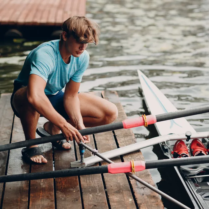 Un jeune homme en t-shirt de sport bleu est assis sur un quai en assemblant des rames pour un bateau, avec de l'eau et un bateau visibles en arrière-plan.