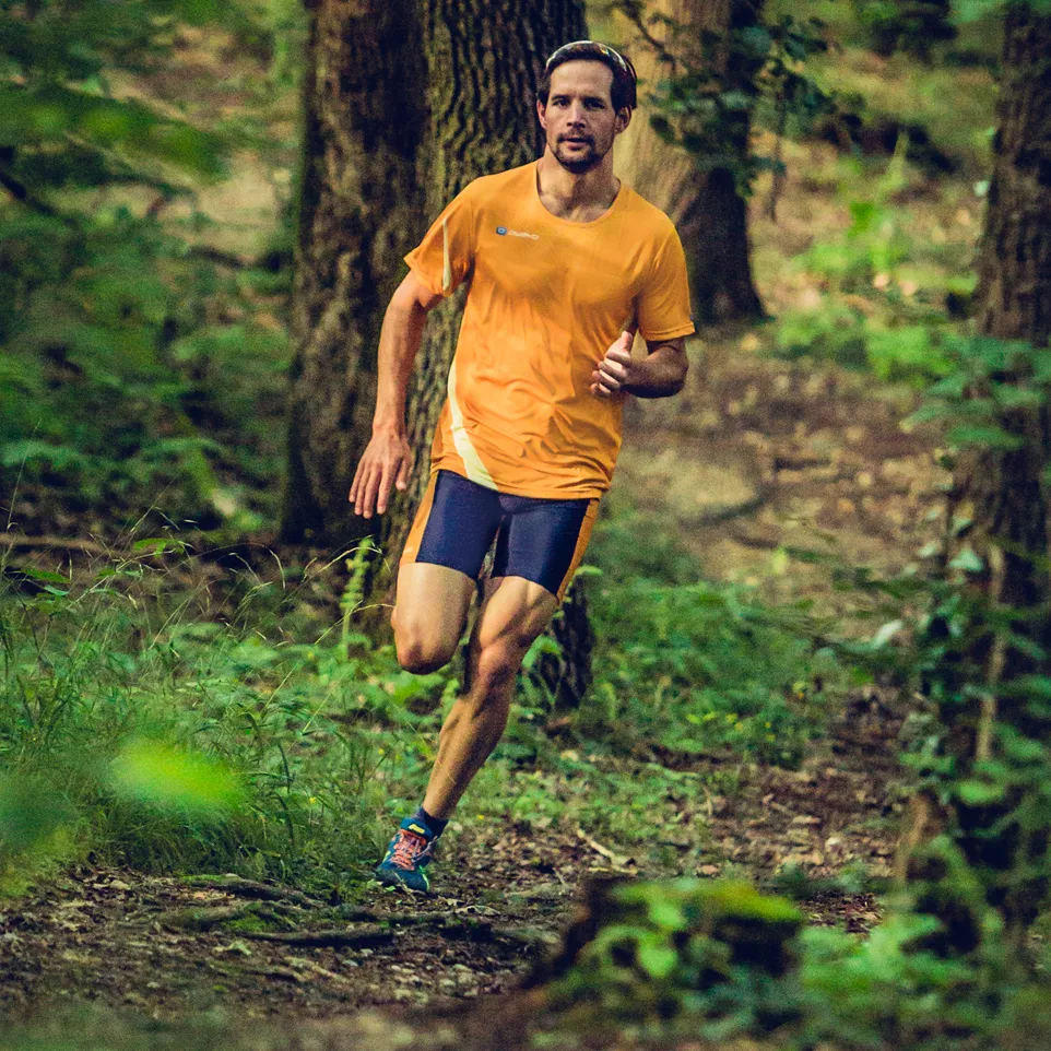 Un homme court sur un sentier forestier portant un maillot de course orange et un short de course bleu foncé.