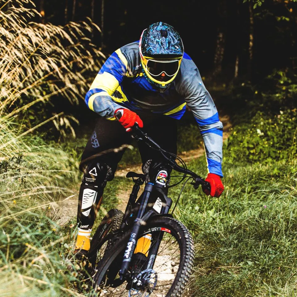 Un cycliste de montagne portant un maillot à manches longues coloré aux accents bleus, jaunes et gris roule sur un sentier forestier.